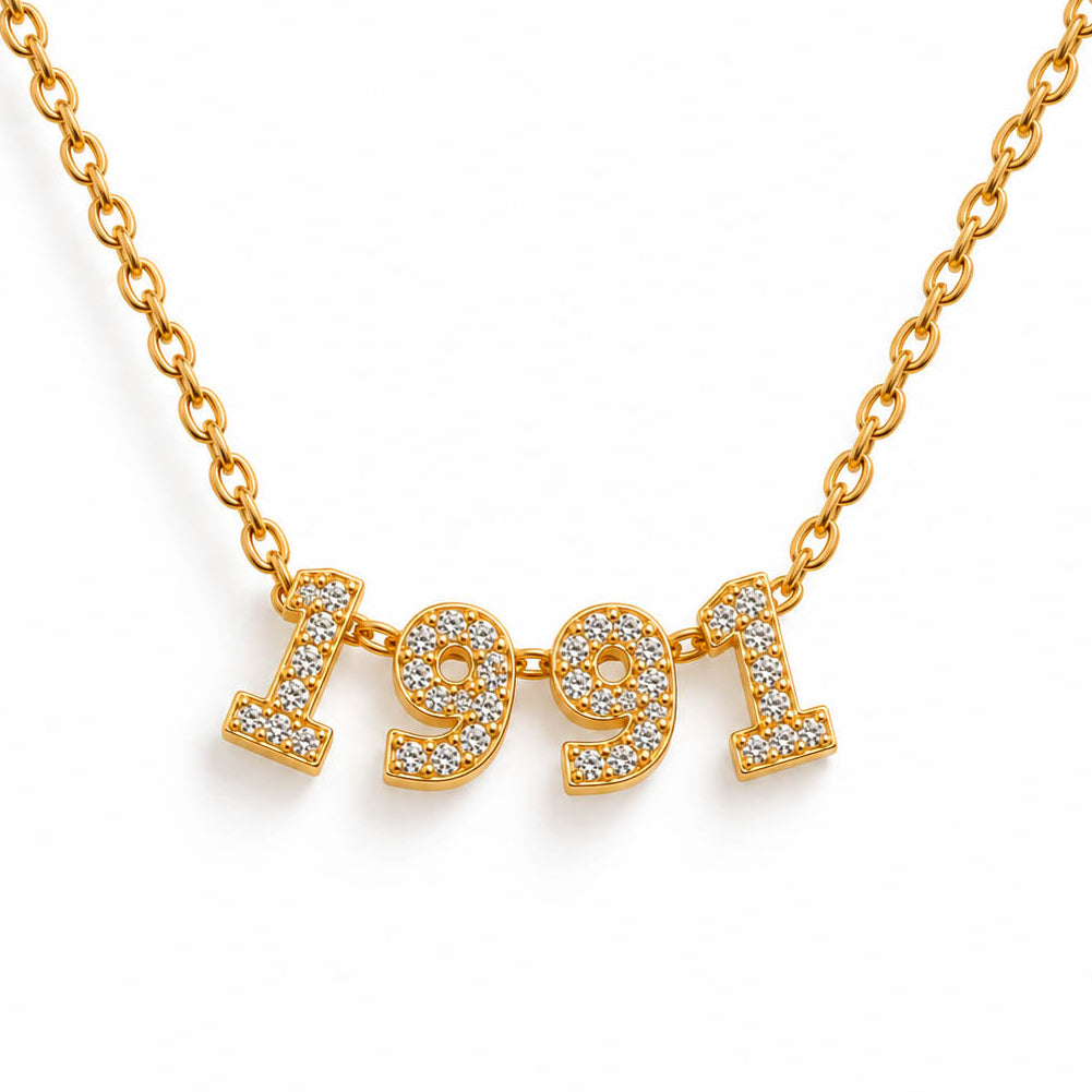 A studio shot of a thin gold link necklace centered with four rhinestone-encrusted charms that spell out the year "1991".