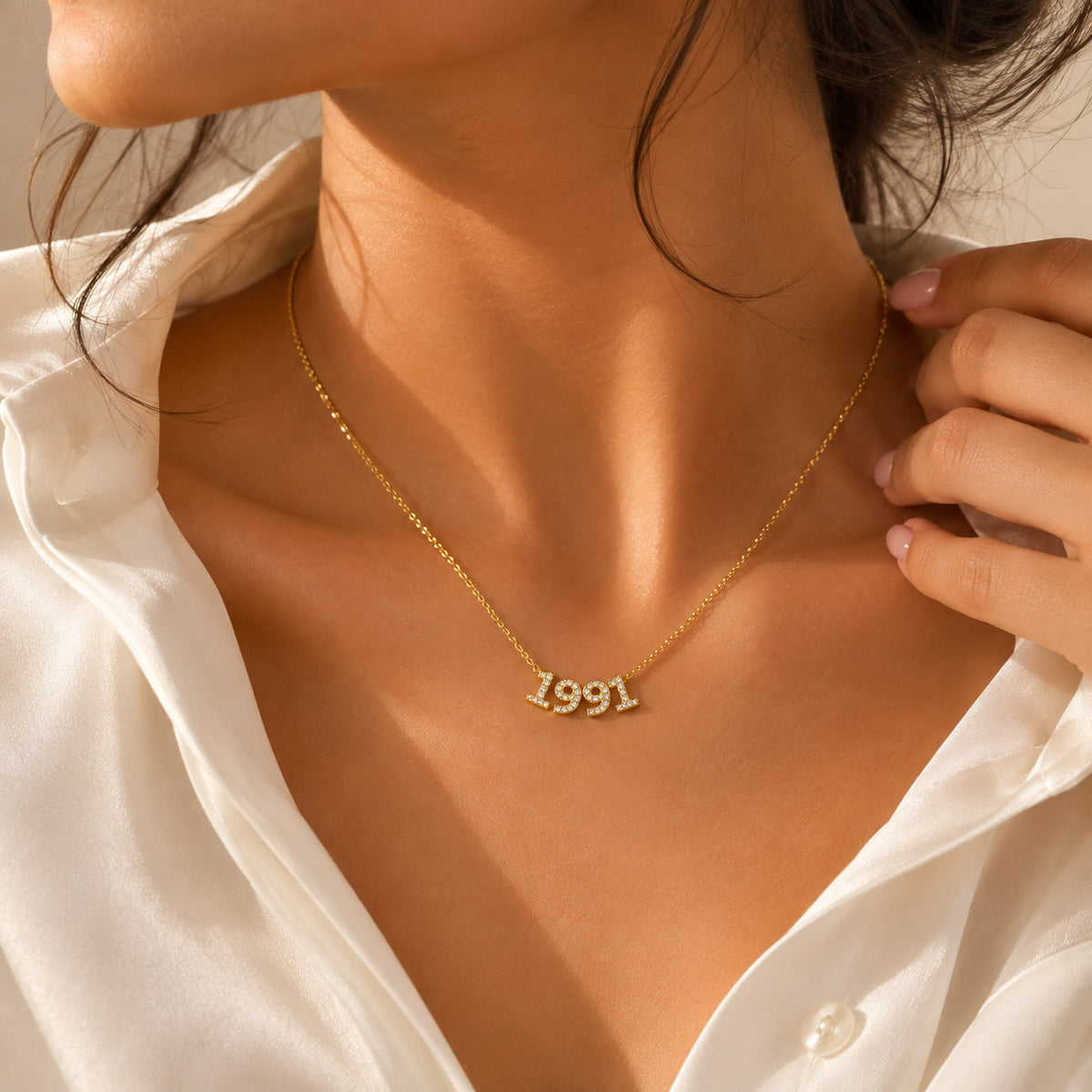 Gold-plated personalized number necklace featuring the sparkling pavé year “1991” on a delicate gold chain, styled on a model wearing a white shirt in warm natural sunlight.