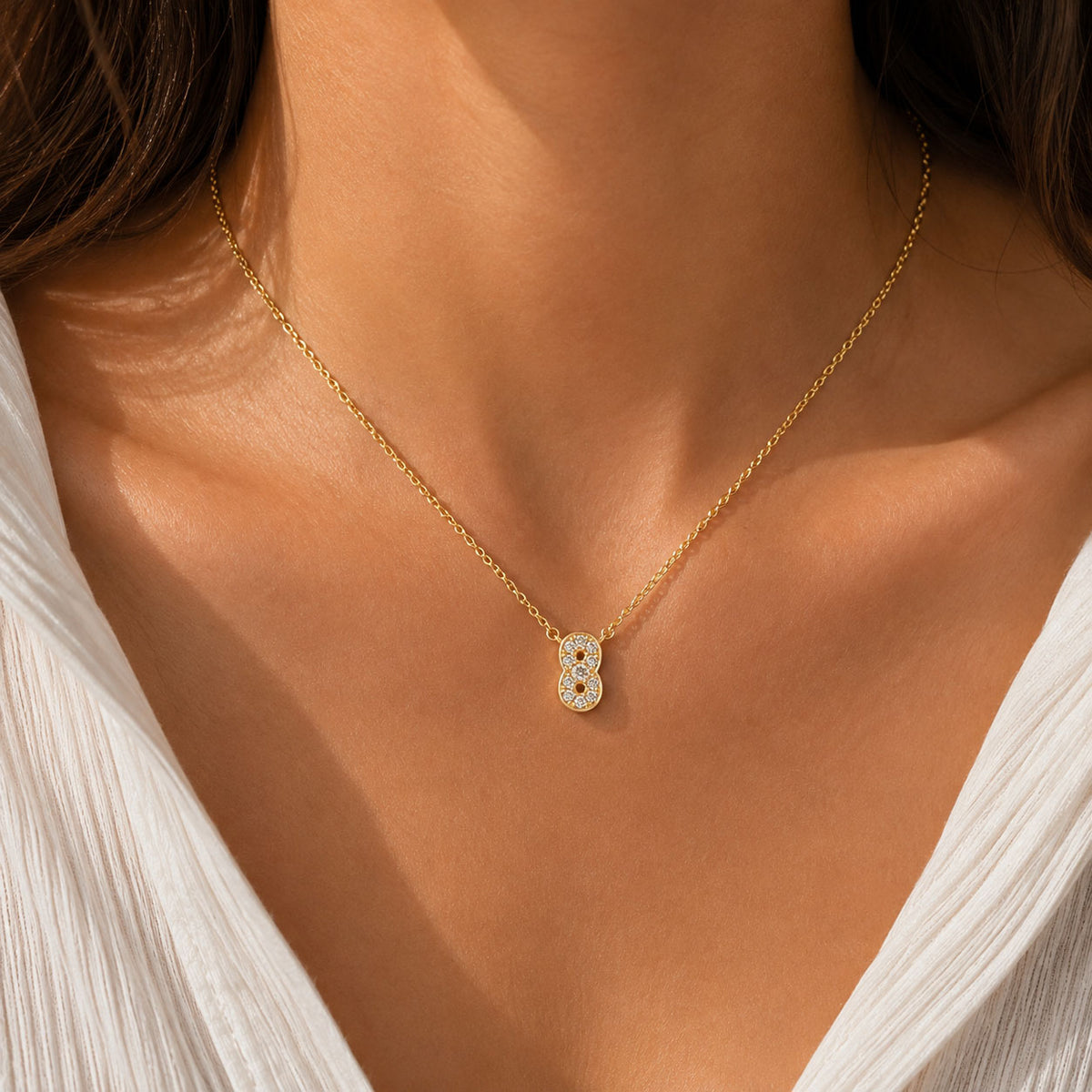 Gold-plated personalized number necklace featuring a sparkling pavé “8” pendant on a delicate gold chain, worn on a model in a white outfit with warm natural sunlight.