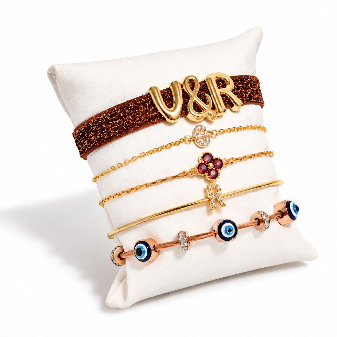 Gold-plated bracelet stack displayed on a white cushion, featuring a brown glitter “V&R” bracelet, delicate chain bracelets with floral and letter charms, a slim gold bangle, and blue Evil Eye bead bracelets.