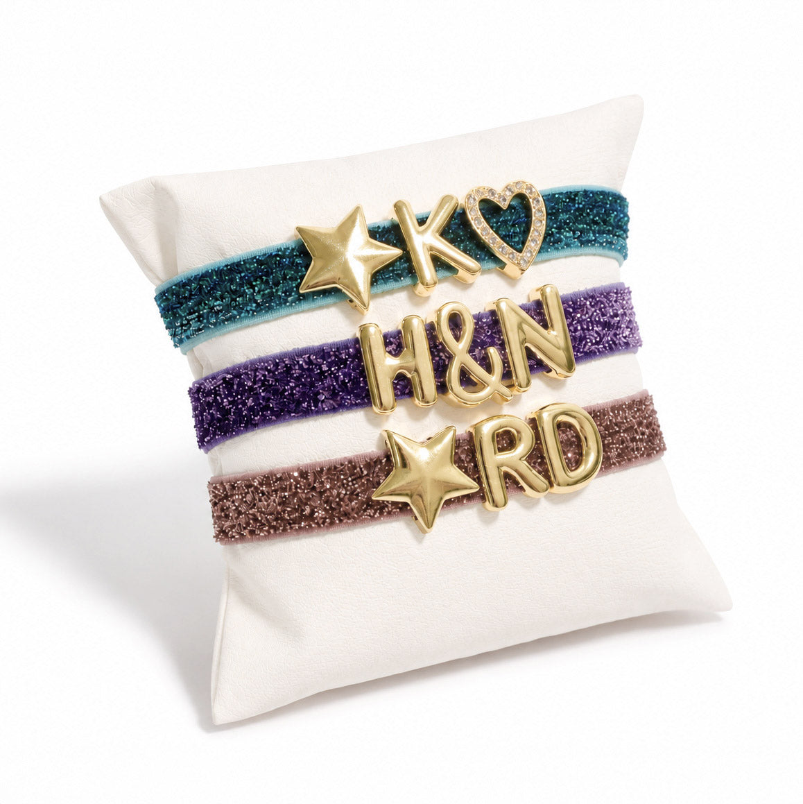 Gold-tone glitter bracelet stack displayed on a white cushion, featuring blue, purple, and brown bands with star and letter charms.