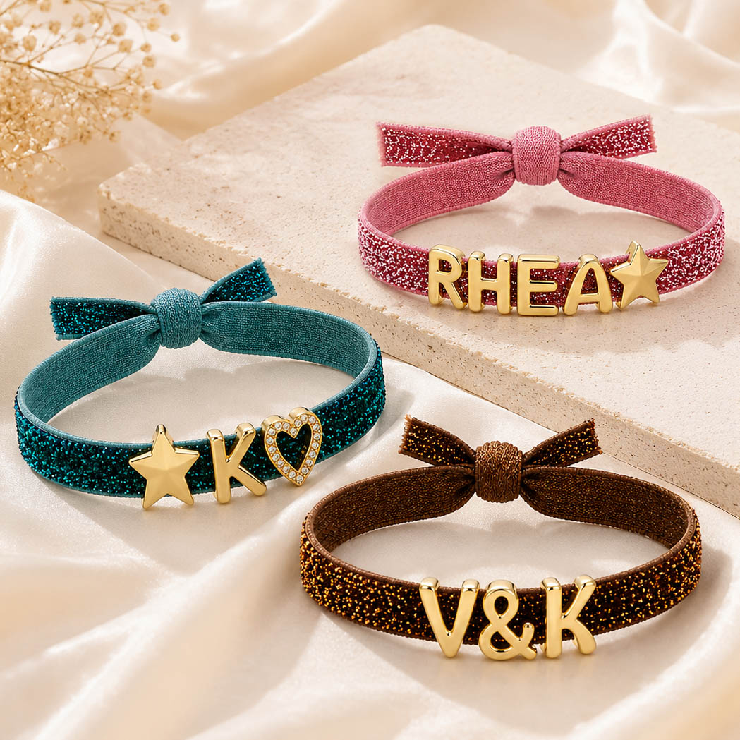 This image shows three glitter bracelets in teal, pink, and brown, styled on satin and stone blocks, with gold initial/heart/star charms.