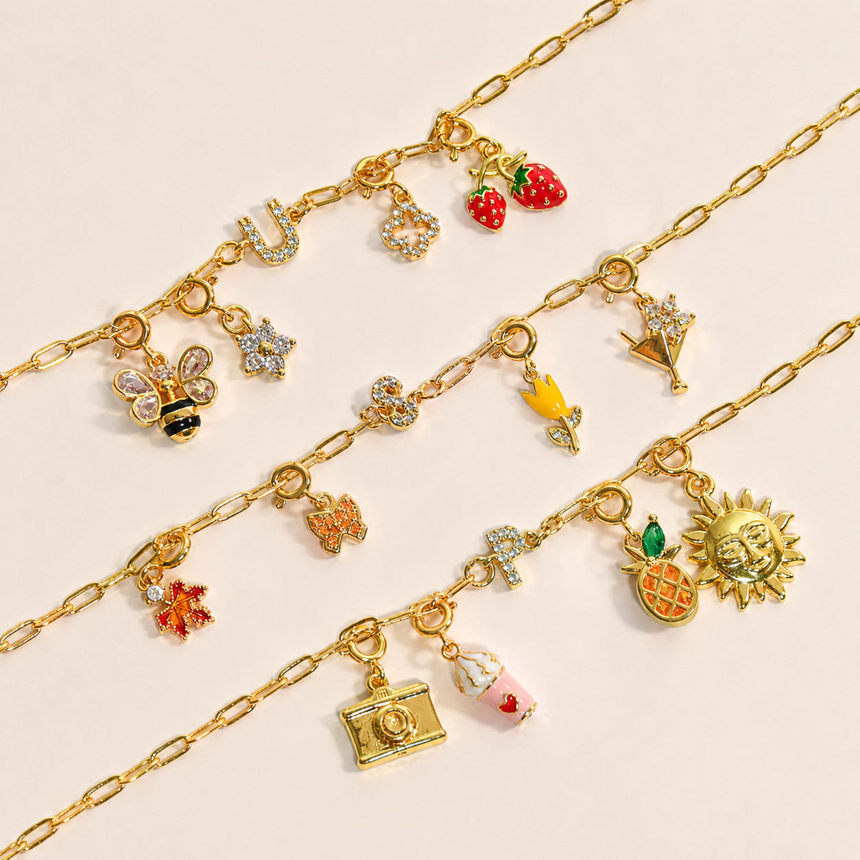 Gold-tone charm necklaces featuring interchangeable charms including strawberries, butterflies, a bee, flowers, stars, a pineapple, sun, camera, and letter charms, arranged on a neutral background.