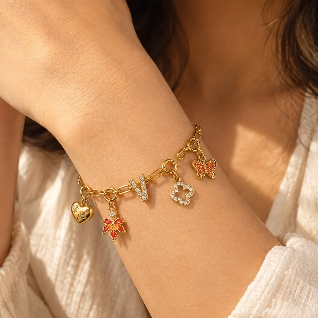 Gold-tone charm bracelet worn on a wrist, featuring a sparkling letter V, heart charm, red maple leaf, orange butterfly, and small decorative charms, styled with a white outfit.