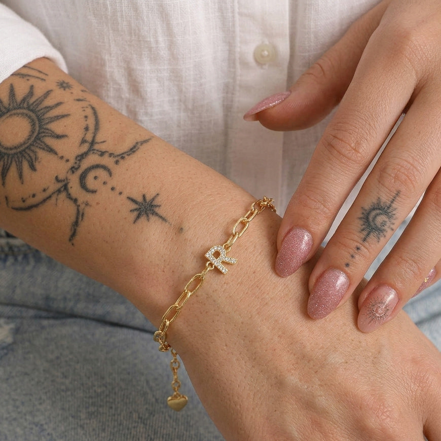 Gold-tone chain bracelet worn on a wrist, featuring a sparkling letter R charm and a small dangling heart charm.