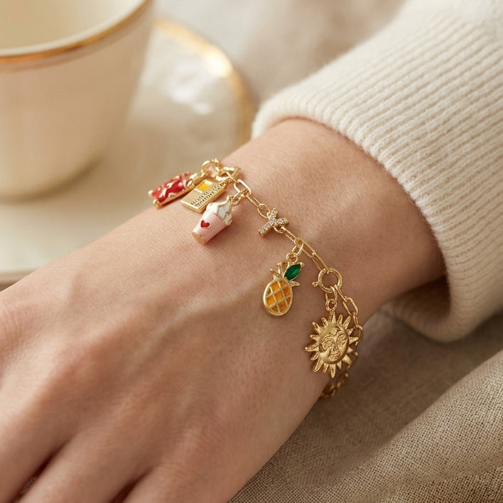 Gold-tone charm bracelet worn on a wrist, featuring a red suitcase, small tag, drink cup, sparkling letter charm, pineapple, and sun charm.