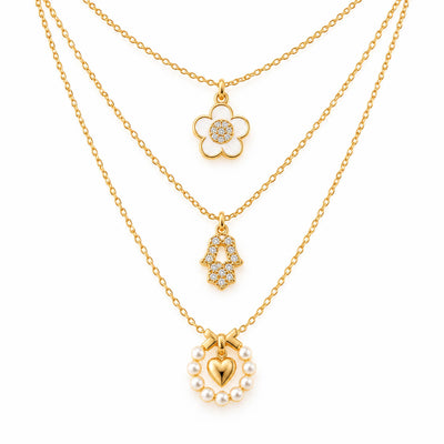 A product photo of a layered gold-tone necklace set featuring a daisy flower pendant with a white enamel outline and crystal center, a rhinestone-paved Hamsa hand charm, and a pearl wreath pendant holding a glossy gold heart beneath a bow.