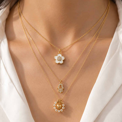 Layered gold necklaces featuring a white flower pendant, sparkling Hamsa pendant, and pearl-accented heart pendant styled on a model.