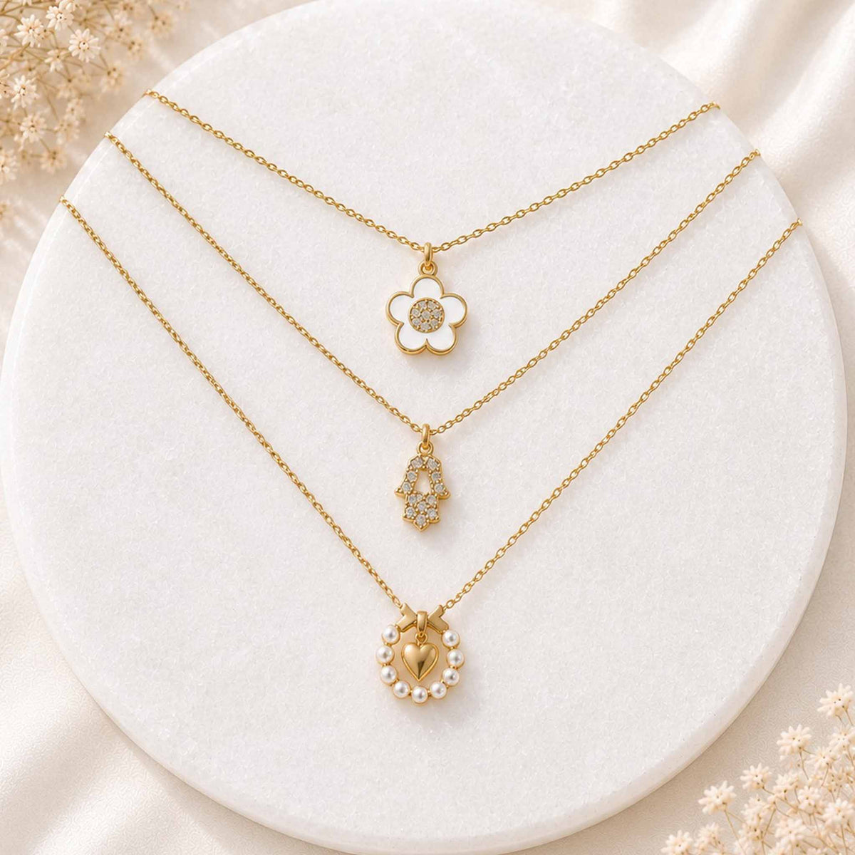 Three delicate gold pendant necklaces featuring flower, Hamsa, and pearl heart designs displayed on a white circular jewellery stand.