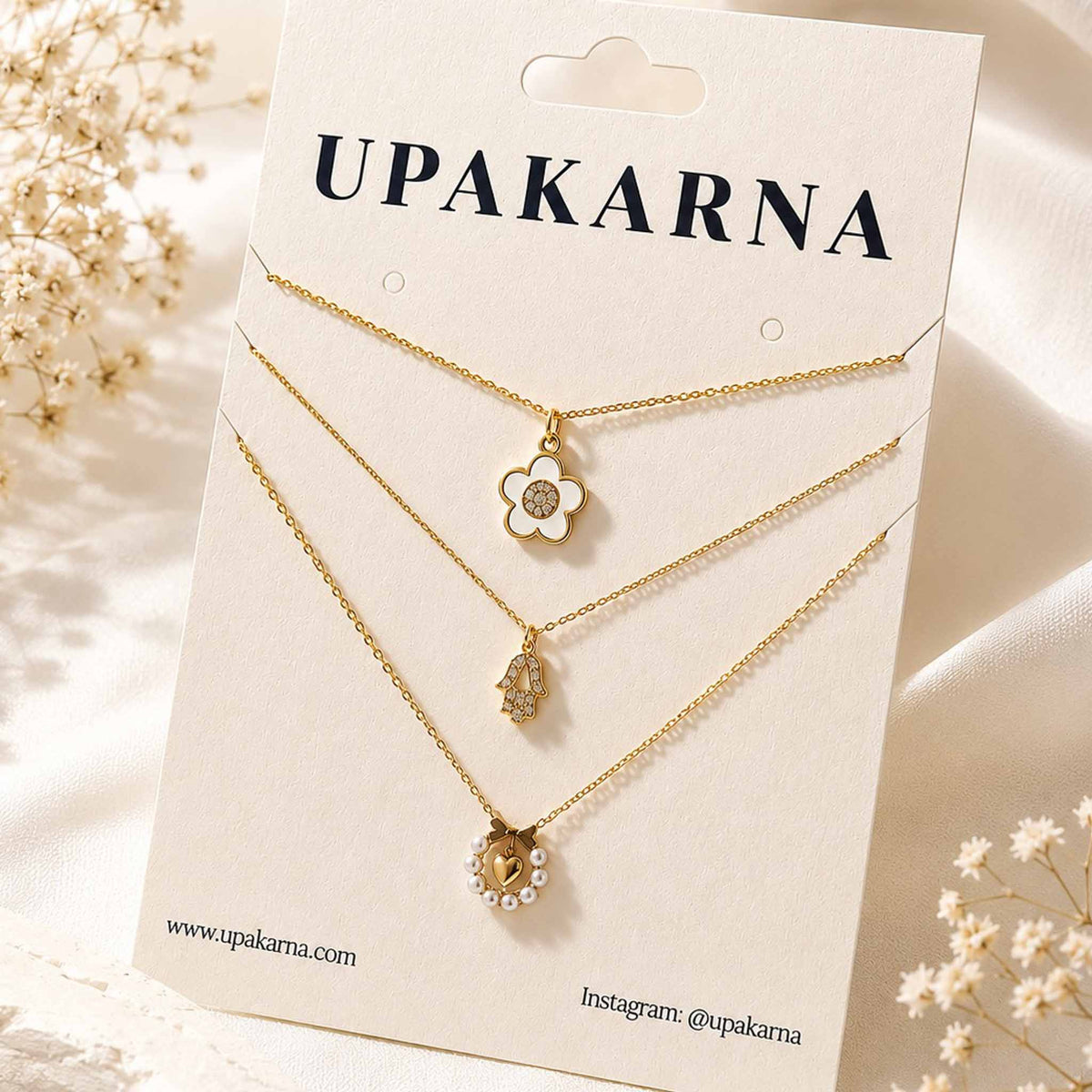 Upakarna layered gold necklace set with flower, Hamsa, and pearl heart pendants displayed on a branded jewellery card.