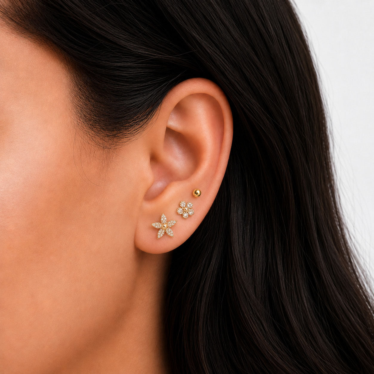 Close-up view of a model’s ear wearing gold-tone floral earrings with clear sparkling stones and pearl details, paired with a small gold ball stud.