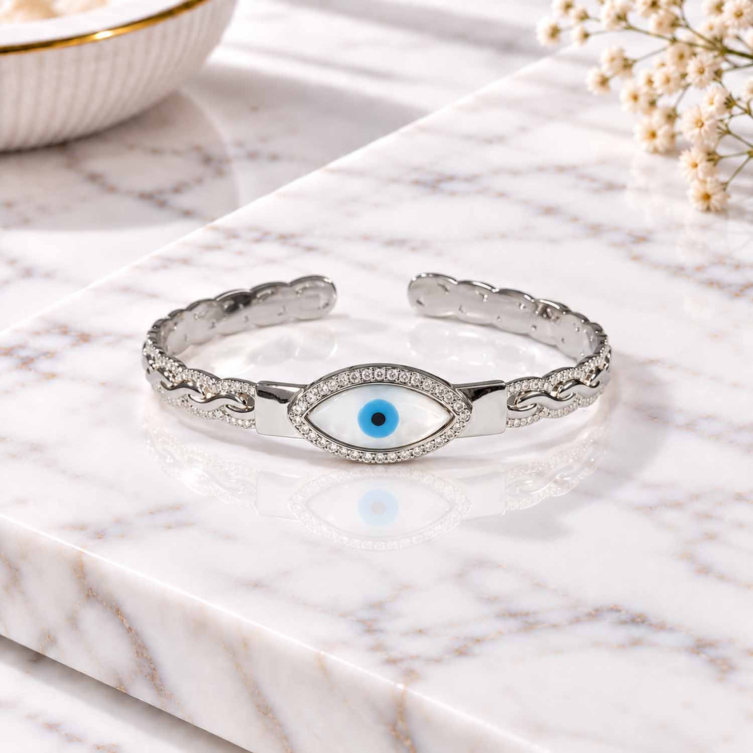 Silver evil eye cuff bangle with crystal detailing and a blue eye centerpiece, displayed on a marble surface.