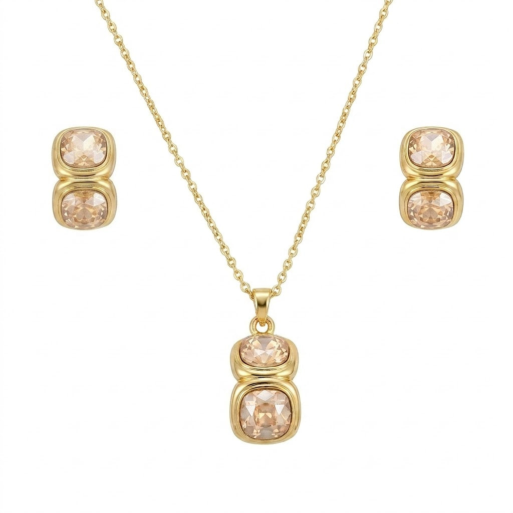 A gold jewelry set isolated on a plain white background, featuring a fine-chain necklace with a drop pendant composed of two vertically stacked, cushion-cut champagne crystals in smooth gold bezel settings. The set is paired with matching stud earrings that replicate the same two-stone stacked design for a cohesive, elegant finish.