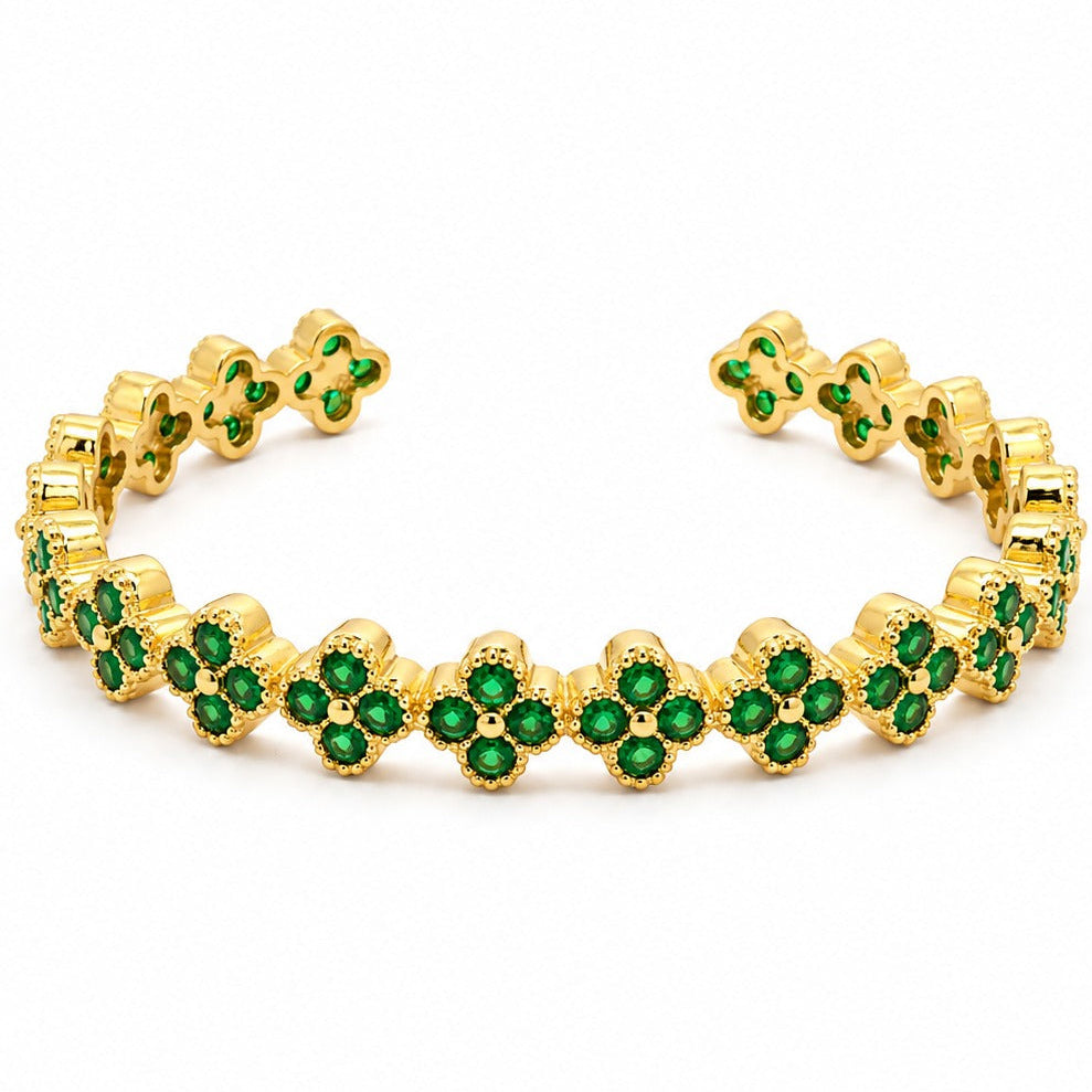 Anti-tarnish yellow gold open cuff bracelet featuring a continuous band of four-leaf clover quatrefoil links, each pave-set with round green emerald cubic zirconia stones and finished with beaded milgrain edges, set against a solid white background.