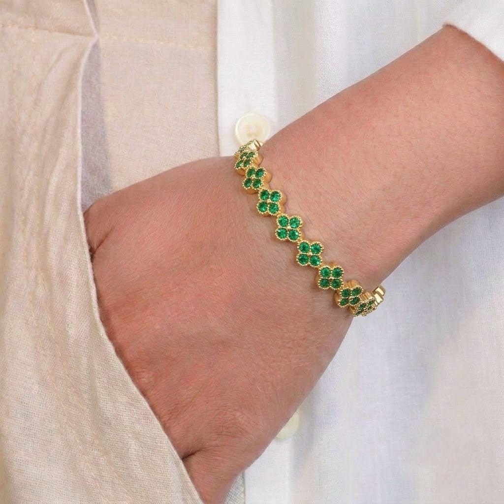 Anti-tarnish yellow gold bracelet styled on a model's wrist, featuring repeating four-leaf clover stations encrusted with vibrant emerald-green cubic zirconia stones and bordered with beaded milgrain edging.