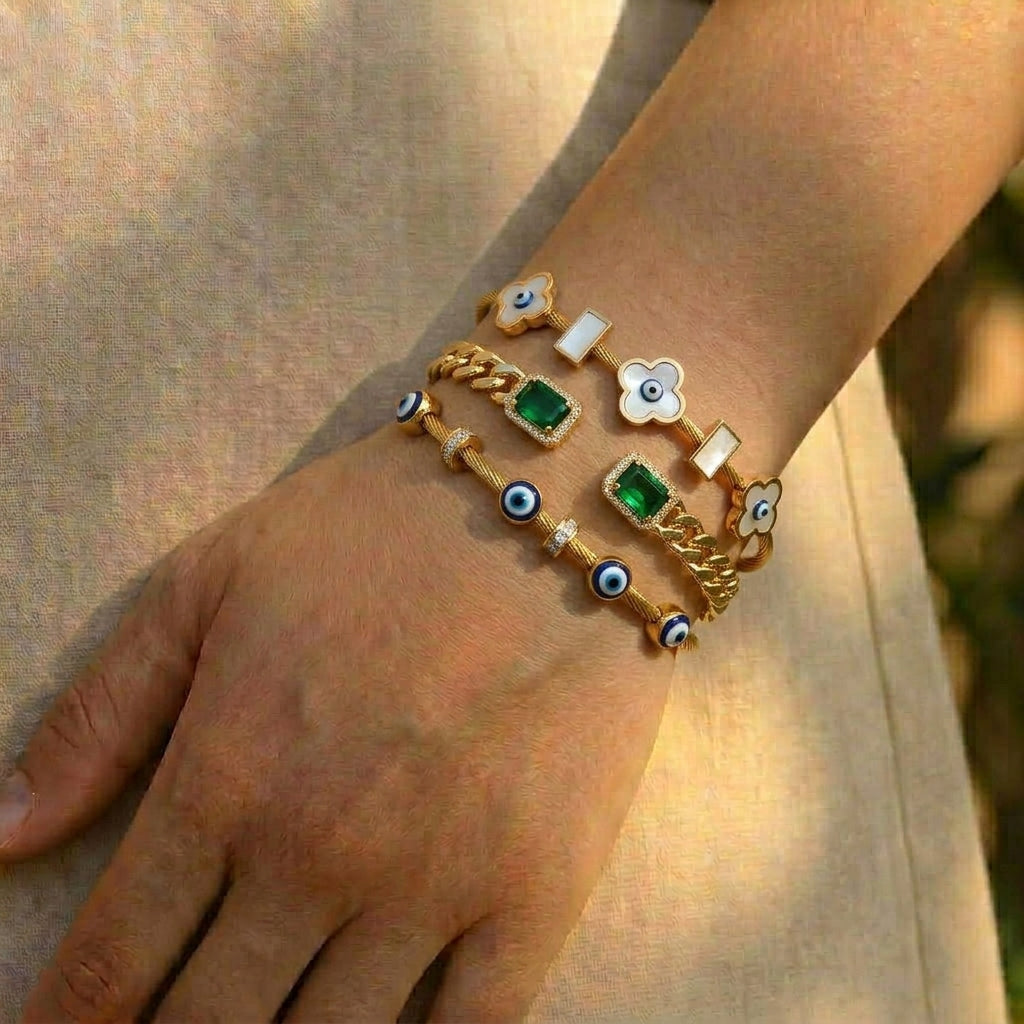 Gold and rose gold anti-tarnish bracelet stack featuring blue evil eye charms, white clover motifs, emerald green gemstones, and crystal accents, elegantly worn on the wrist in natural sunlight.