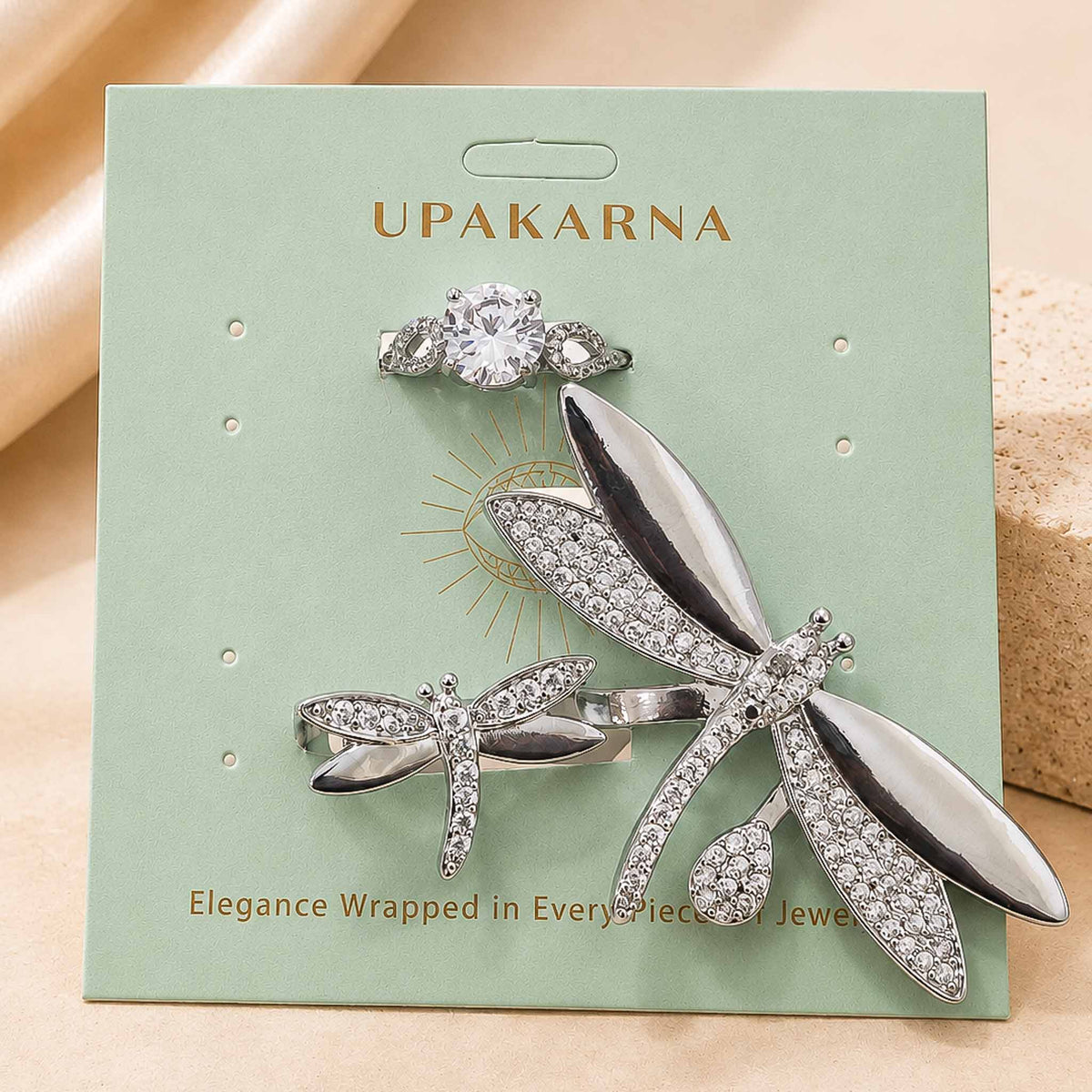 Upakarna jewellery display featuring sparkling silver-toned dragonfly jewellery and a crystal stone ring on a sage green branded card.