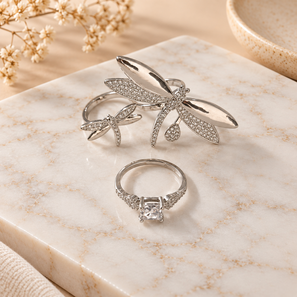 Two silver-tone rings arranged on a white marble surface, featuring polished metal designs with crystal embellishments and intricate decorative details, styled with soft floral accents.