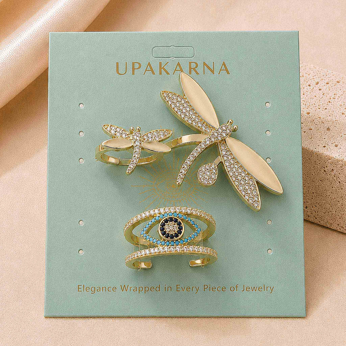 Upakarna jewellery display card featuring elegant gold-toned dragonfly and evil eye jewellery designs with sparkling stone details on a soft sage green background.