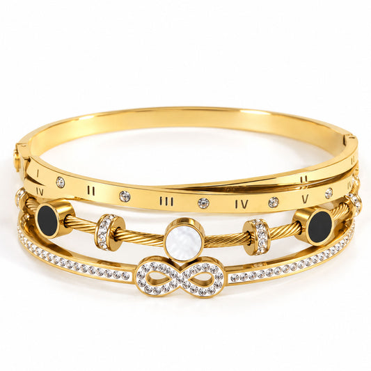 A product photo of a stacked set of gold-tone bracelets featuring Roman numeral markers, a twisted cable wire band with black and mother-of-pearl stations, and a crystal-accented band centered with a double infinity symbol.