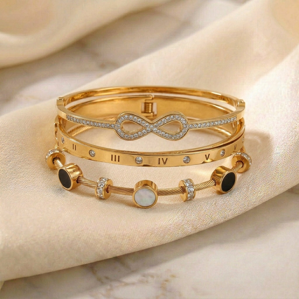 Luxury gold anti-tarnish bracelet stack featuring a crystal infinity bangle, Roman numeral bangle, and a mother-of-pearl cable bracelet with sparkling crystal accents, elegantly displayed on soft satin fabric.