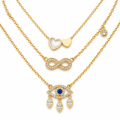 Upakarna gold necklace set featuring three layered necklaces with a heart pendant, crystal infinity pendant, and evil eye pendant with dangling crystals on a clean white background.