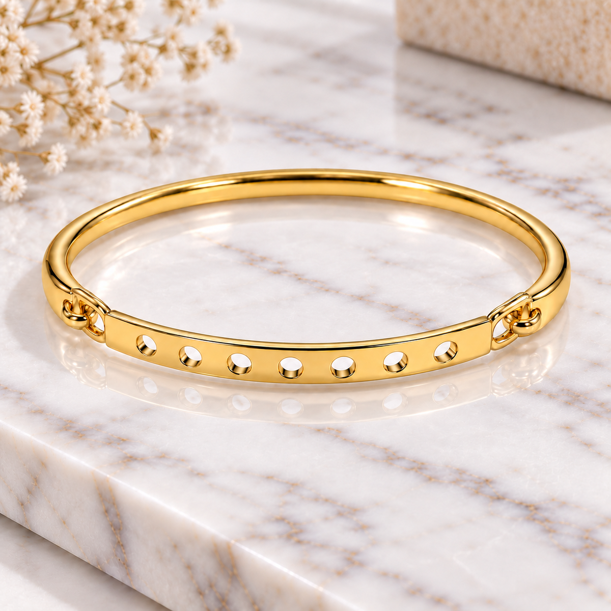 Gold-tone bangle bracelet with six circular cutouts displayed on a white marble surface with soft floral decor.