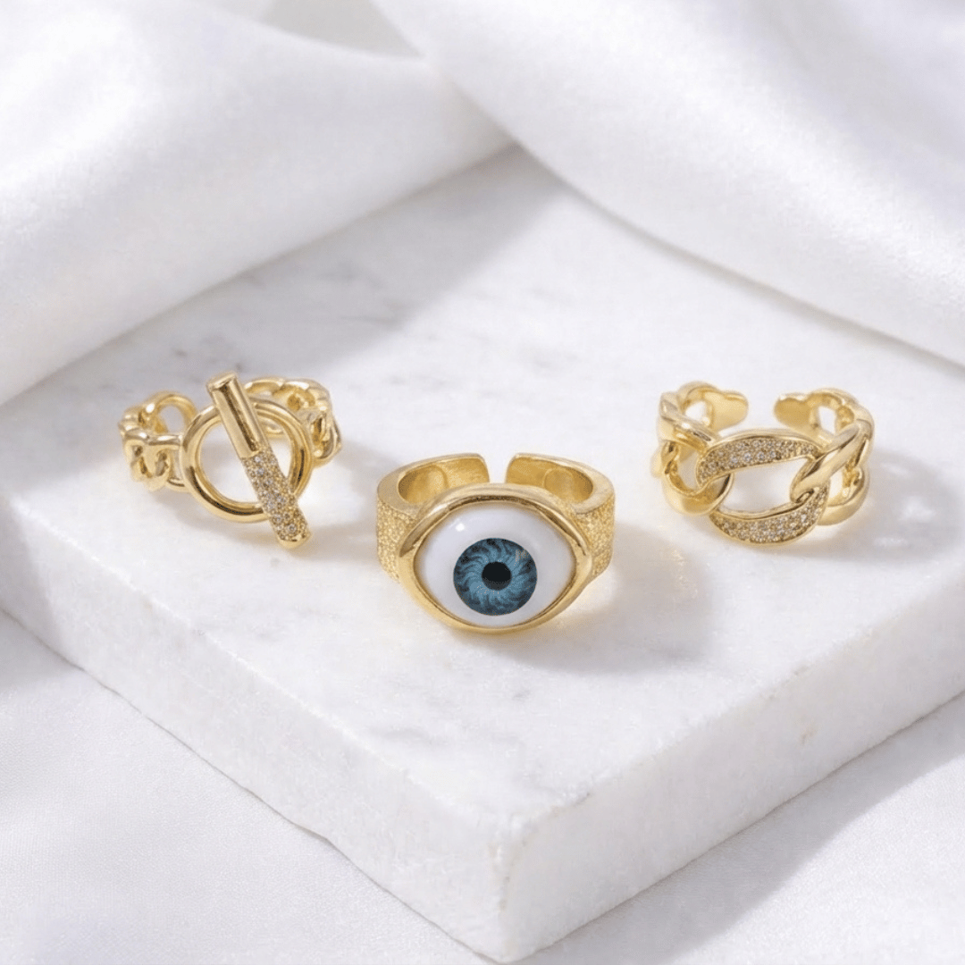 A stack of three adjustable gold-toned rings: a sculptural spike spiral, a chunky chain-link band, and a central signet ring featuring an evil eye symbol.