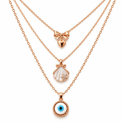 A product photo of a layered rose gold-tone necklace set featuring a bow pendant with a hanging heart, a seashell pendant with a tiny crystal starfish, and a circular evil eye amulet.   