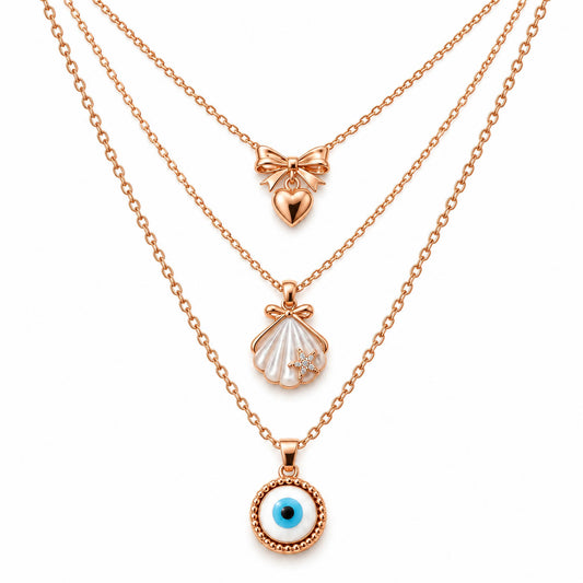 A product photo of a layered rose gold-tone necklace set featuring a bow pendant with a hanging heart, a seashell pendant with a tiny crystal starfish, and a circular evil eye amulet.   