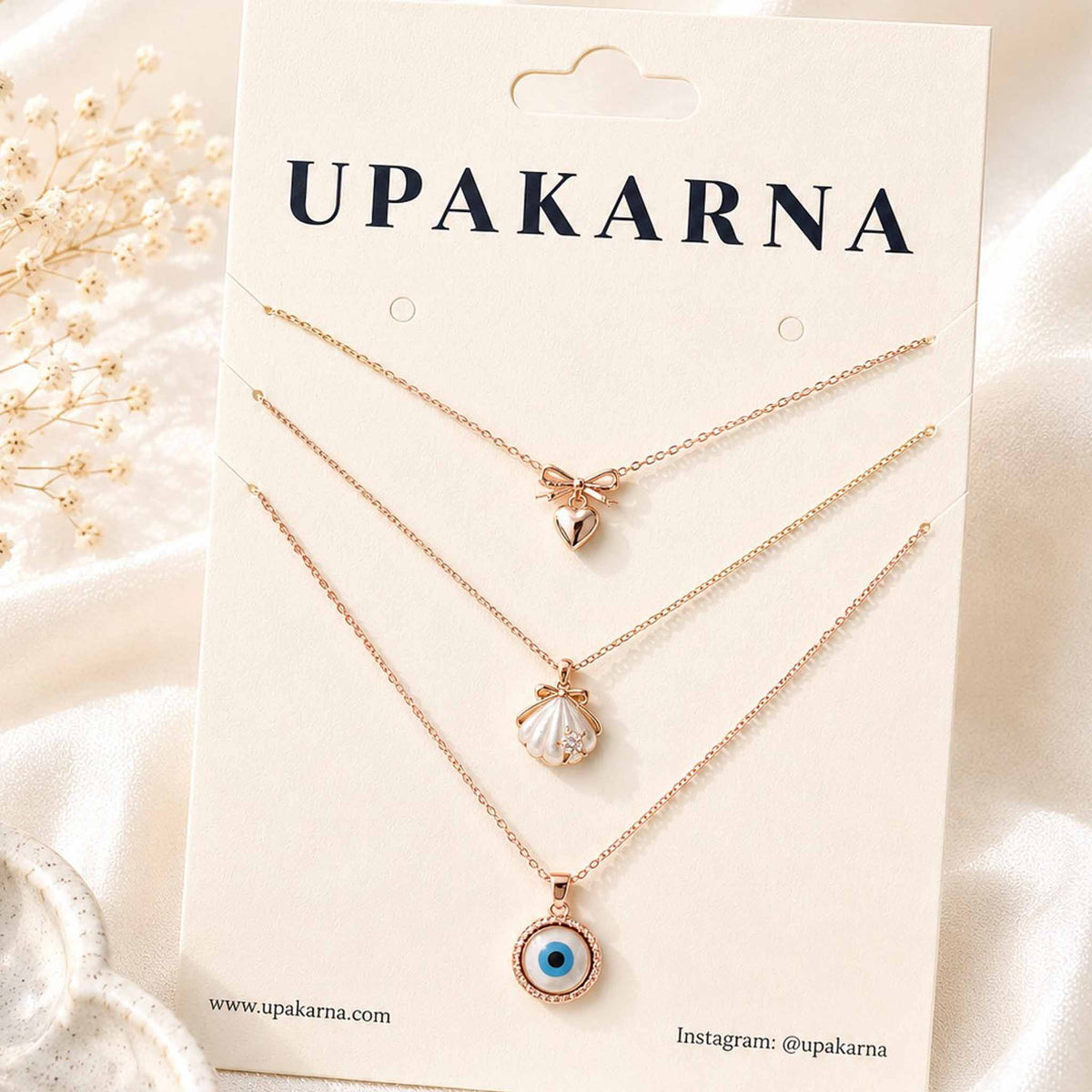 Gold layered necklace set featuring a bow heart pendant, pearl shell pendant, and blue evil eye pendant displayed on an Upakarna jewellery card.