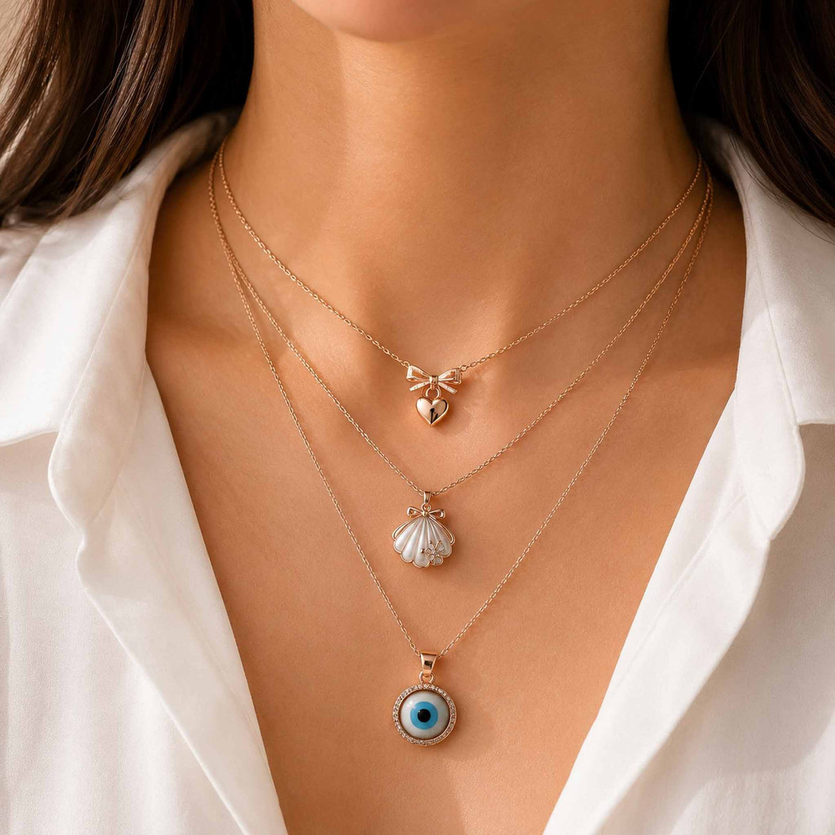 Gold layered necklace set featuring bow heart, pearl shell, and blue evil eye pendants worn with a white outfit.