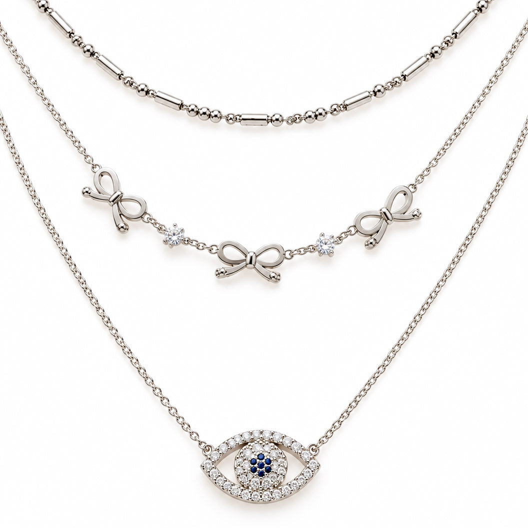 A product photo of a layered silver-tone necklace set featuring a beaded bar chain, a link chain decorated with alternating ribbon bows and round crystals, and a pendant shaped like an evil eye paved in clear crystals with dark blue center stones.