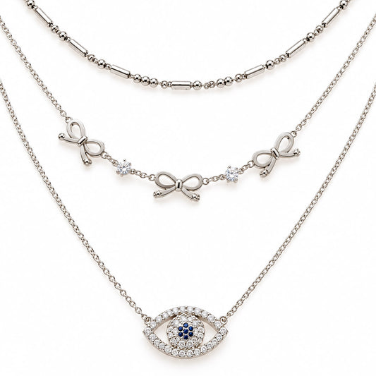 A product photo of a layered silver-tone necklace set featuring a beaded bar chain, a link chain decorated with alternating ribbon bows and round crystals, and a pendant shaped like an evil eye paved in clear crystals with dark blue center stones.