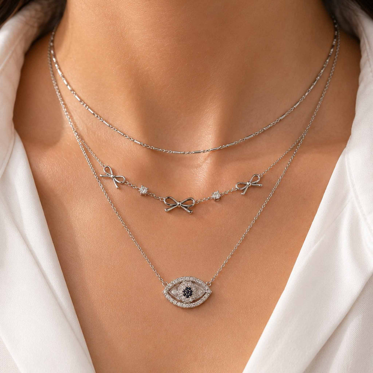 Silver layered necklace set featuring delicate bow charms and a sparkling evil eye pendant, worn with a white outfit.