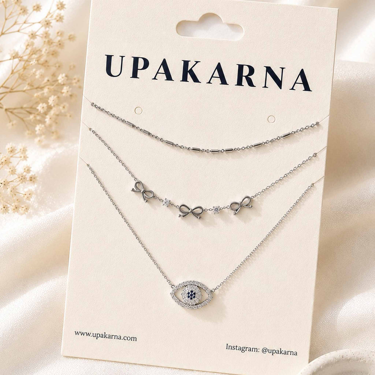 Silver bow charm and evil eye layered necklace set displayed on an Upakarna jewellery card.
