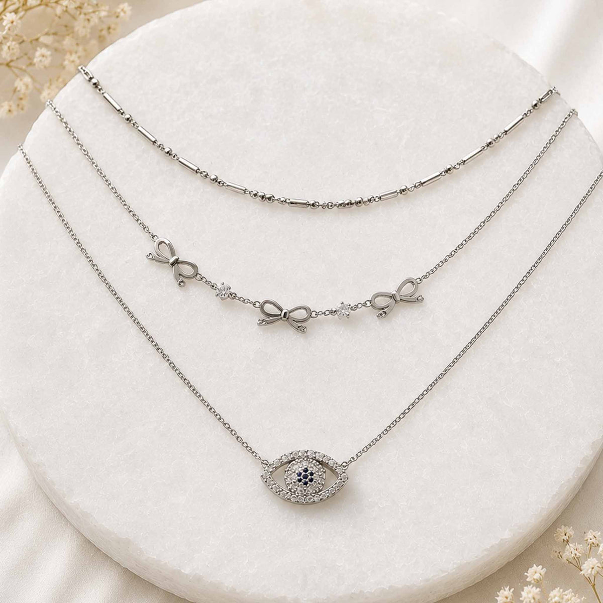 Silver layered necklace set with bow charms and crystal evil eye pendant displayed on a white circular jewellery stand.