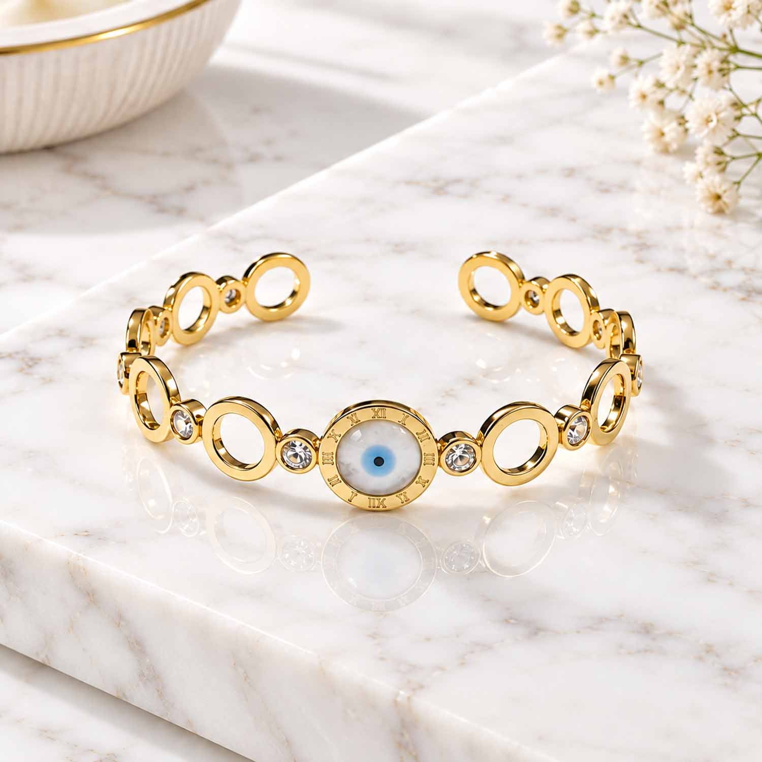 Gold bracelet featuring a central blue evil eye charm surrounded by Roman numeral detailing, paired with polished oval links and sparkling crystal accents, displayed on a marble surface with soft natural lighting.
