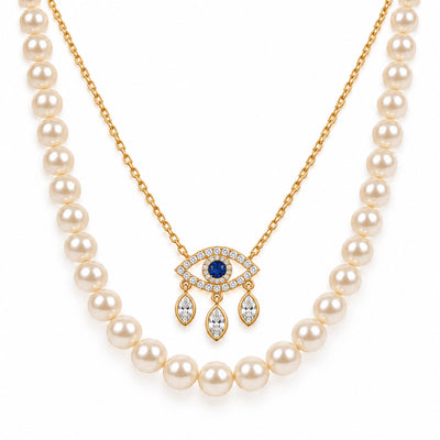 A product photo of a layered gold-tone necklace set featuring a classic white pearl strand paired with a delicate link chain holding an evil eye pendant adorned with crystal pave and dangling marquise stones.   