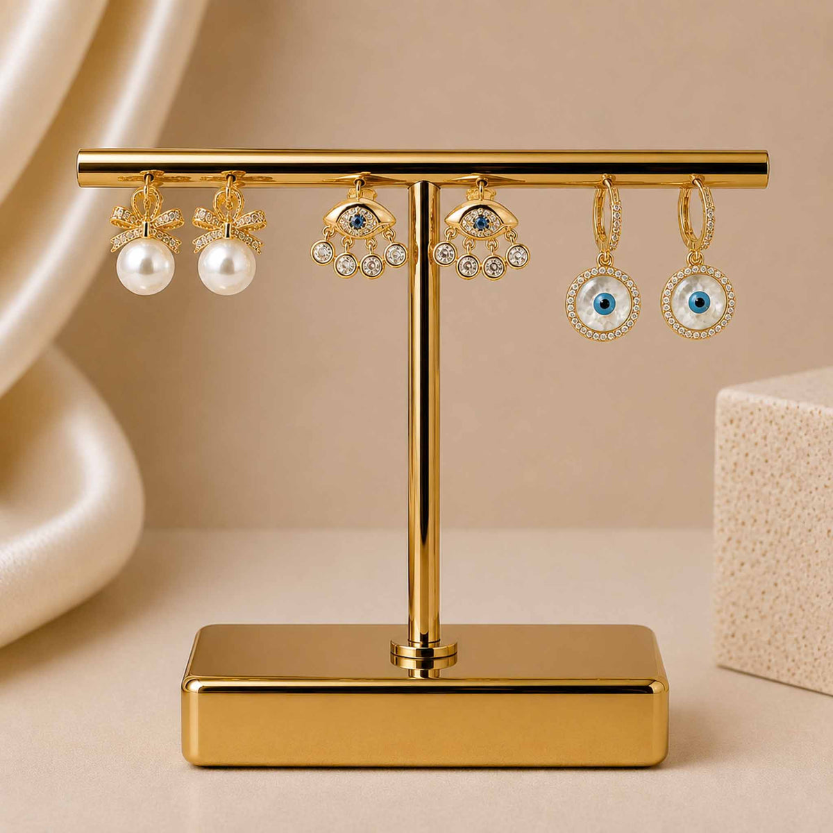 Gold-tone earring stack featuring pearl bow earrings, crystal evil eye dangler earrings, and evil eye hoop earrings displayed on a gold jewellery stand against a soft beige background.