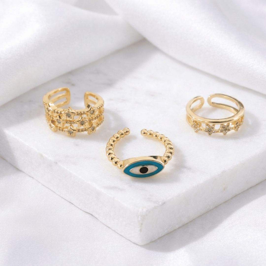 A trio of adjustable gold-toned rings stacked together, featuring a pastel evil eye centerpiece, a sparkling stardust band, and a delicate star-inspired ring.