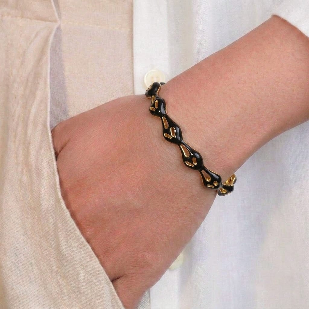 A close-up of a person’s wrist wearing a sleek black enamel bangle with an organic, wavy design and gold-toned accents peeking through the pattern. The bracelet is styled against a neutral beige outfit, creating a modern and minimal contrast.