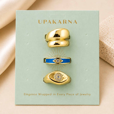Upakarna jewellery display featuring three gold-toned rings, including bold dome rings and blue evil eye designs, arranged on a sage green branded card.