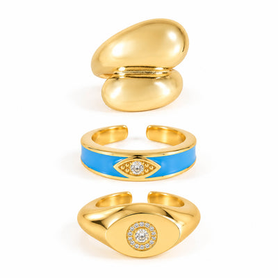A product photo of three gold-tone adjustable rings featuring a chunky organic dome design, a vibrant blue enamel band with a crystal evil eye motif, and a signet-style band centered with a round crystal pave cluster.