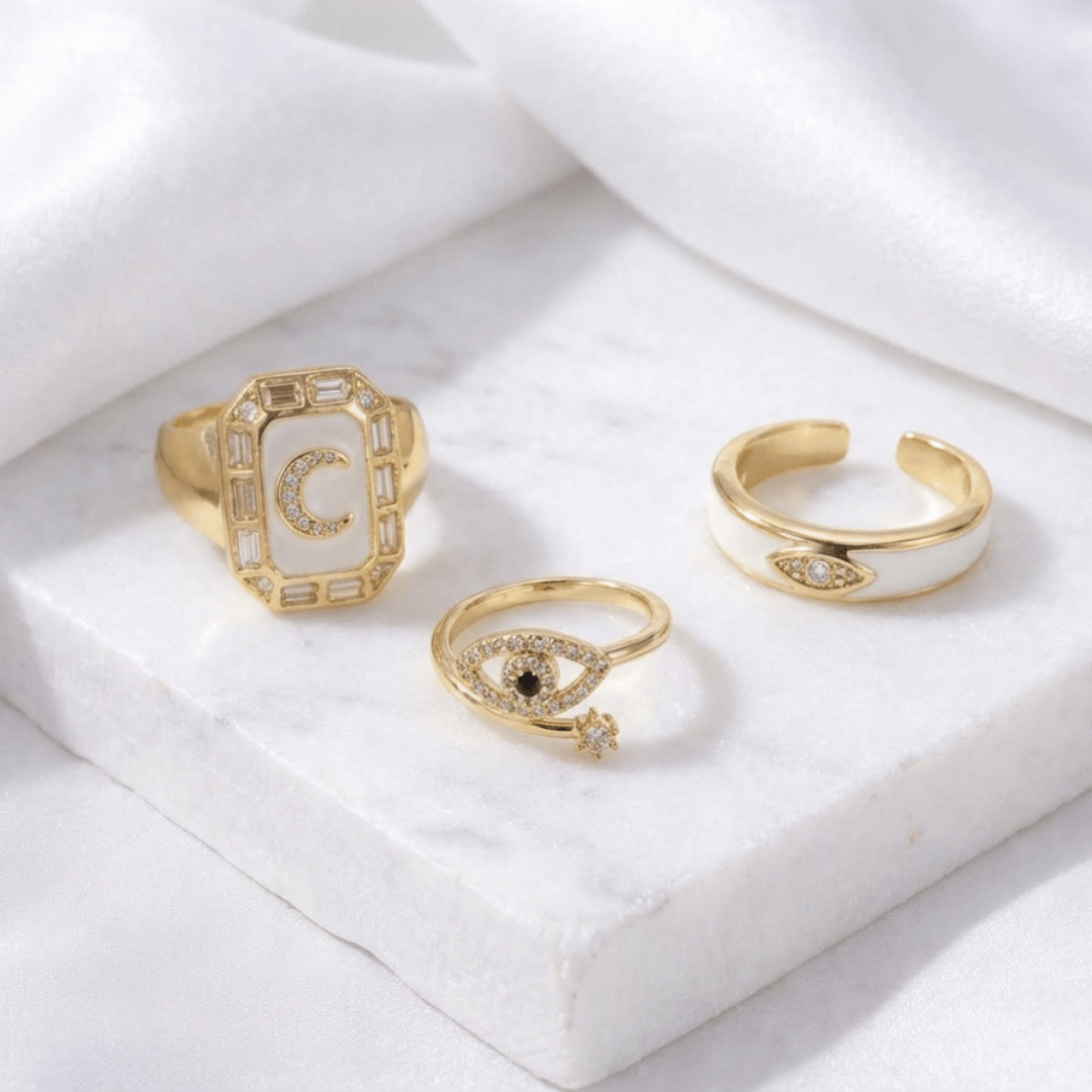 A gold-toned ring stack featuring a blue moonbeam ring, an ivory enamel mystic evil eye, and a crystal-encrusted blink motif.