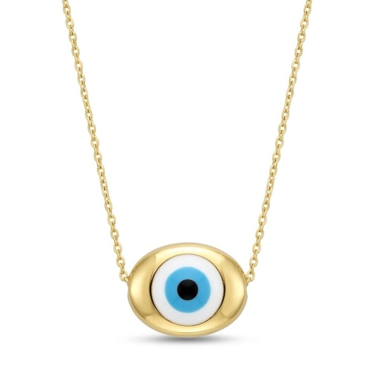 A gold necklace with a horizontally oriented oval charm attached directly to a fine chain. The smooth gold setting frames a classic Evil Eye motif with a black pupil, light blue iris, and white ring, centered on a plain white square background with a dark border.