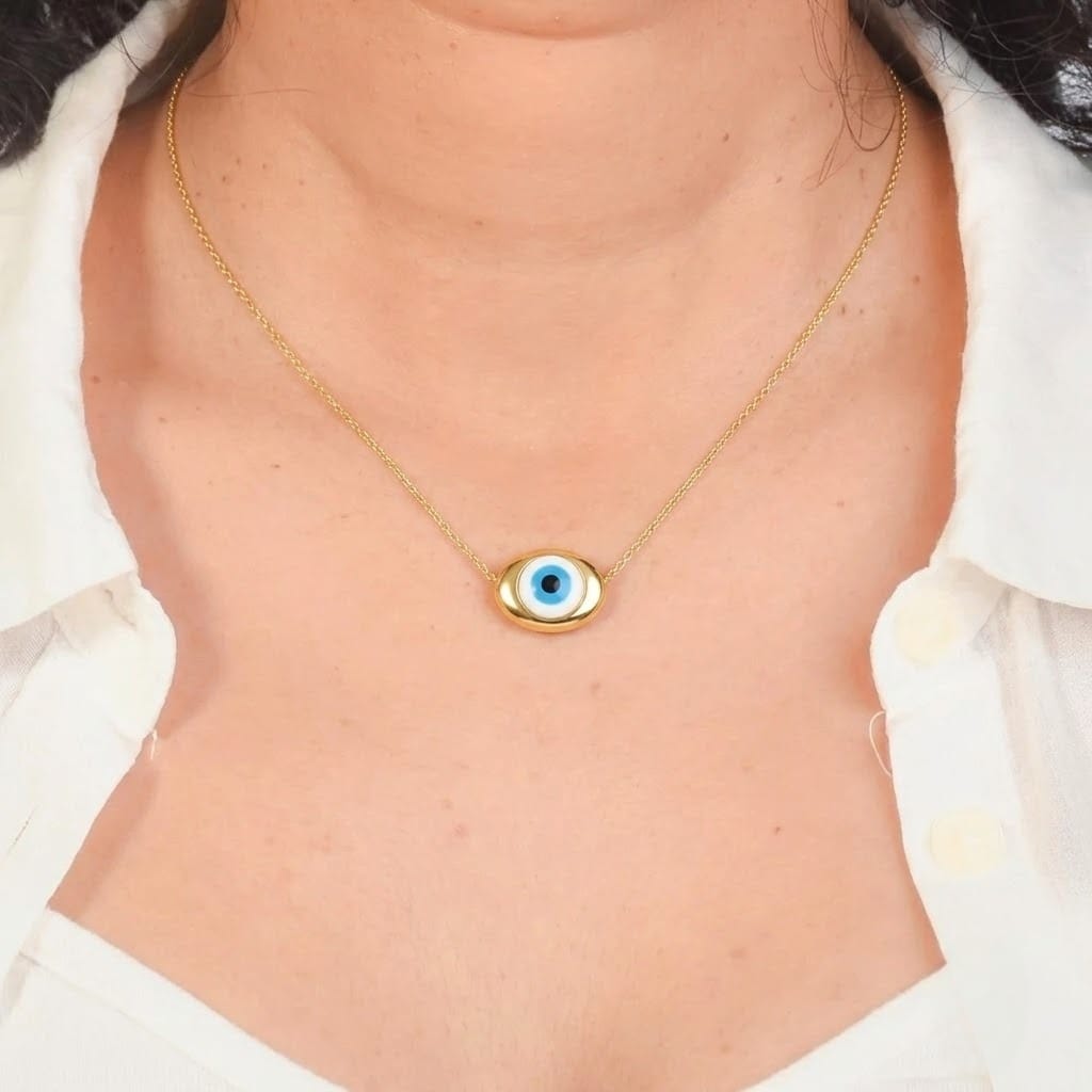Close-up view of a model wearing a fine gold chain featuring a smooth, thick gold oval charm with a central light blue and black Evil Eye design. The necklace rests on the chest framed by an open white collared shirt, presented against a dark gray background border.
