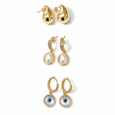 Gold-tone earring stack featuring polished teardrop stud earrings, oval pearl dangler earrings with crystal-studded tops, and circular blue evil eye hoop earrings with crystal detailing, displayed on a clean white background.