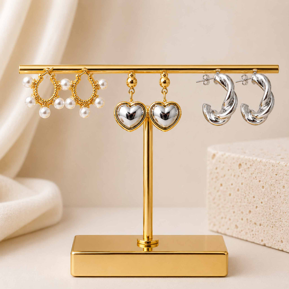 Gold, silver and pearl earring stack featuring pearl-detailed hoops, two-tone heart drop earrings and twisted silver hoops, displayed on a gold jewellery stand with a beige background.