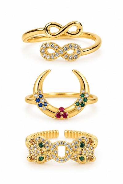 Three gold statement rings featuring an infinity duo ring, a colorful crescent crystal ring, and a bold butterfly-inspired pavé ring with green gemstone accents on a clean white background.